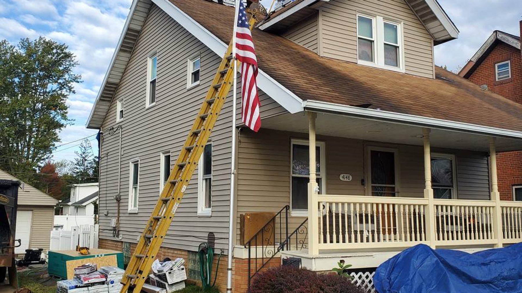 A Stormfront roof job in progress: a ladder up the side of a porch house under an American flag, shingle bundles stacked below, and a blue tarp by the porch