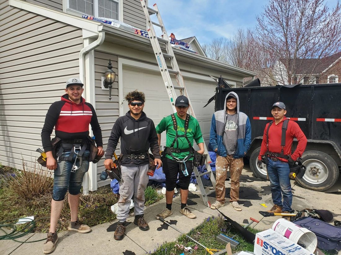 Five Stormfront crew members in tool belts on a driveway, with a ladder to the roof and a dump trailer behind them